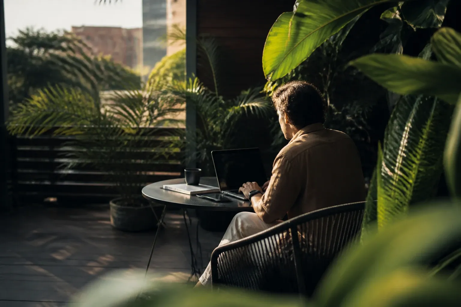Persona trabajando con una laptop en una terraza rodeada de plantas tropicales, con luz natural del atardecer